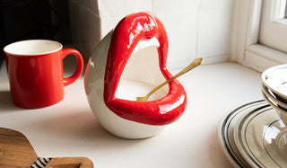 A salt pig in the shape of lips with a spoon inside, styled with a red mug, a wooden serving board and some plates and bowls, styled on a window ledge in front of a window.