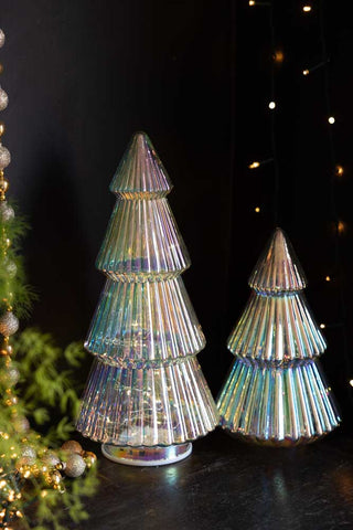 The Large Glass Christmas Tree With LED Lights displayed next to a smaller version in front of a dark wall, styled with some greenery, beads and fairy lights.