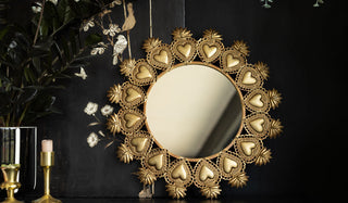 The Gold Milagro Heart Mirror leaning against a black wall, styled with greenery and candlestick holders.