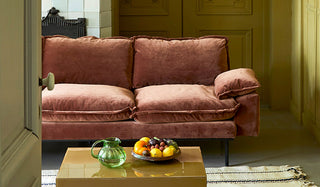 The HKliving Retro Sofa in Magnolia Velvet styled in a living room behind a rug and coffee table, with a fruit bowl and glass jug.