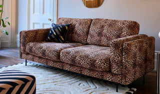 The Grace Medium Sofa In Leopard Love Velvet Natural styled in a living room with a cushion, rug, footstool and plant.