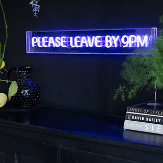 The Please Leave By 9pm Neon Lightbox displayed on the wall in a dark room, styled with plants and books.