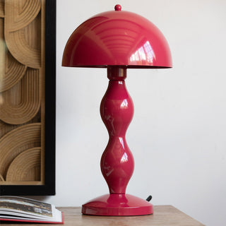 The Raspberry Red Wavy Retro Table Lamp displayed on a wooden surface, styled with an open book and an art print on the wall.