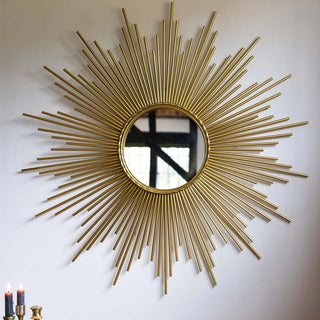 A round gold starburst mirror on a white wall, above some lit candles.