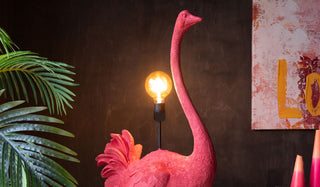 A Large Pink Ostrich Floor Flamp in front of a dark wall, styled with a plant, canvas and candles.