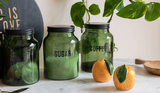 Three green kitchen tea/coffee/sugar storage jars displayed on a marble surface with orange salt and pepper shakers, a spoon and serveware, with a plant in the background.