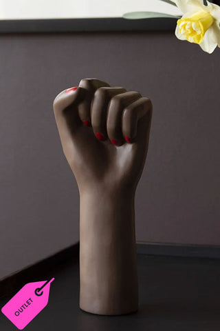 The black girl power vase in a dark and moody room. The vase is emblemetic of a womans fist power pose. In the bottom left hand side is the label outlet.