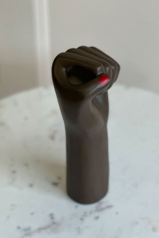 A black womans hand in a fist vase with red nails on a white marble table.