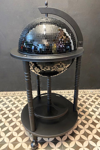 An all-black disco ball drinks trolley with a black base with wine holders. The trolley is on wheels and is styled on a tile floor with a black background.