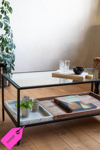 The rectangular glass table in a living room. The top is glass and the bottom shelf has a marble and a wood tray on it; perfect for trinkets and living room essentials. In the bottom left-hand corner is the text: outlet.