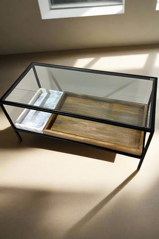 A beautiful glass coffee table up in a bare natural light filled room. The frame of the table is black and on the bottom shelf is a large wooden tray and a smaller white marble tray. The background is neutral.