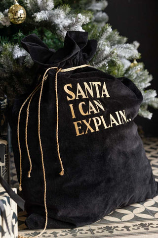 Black and gold velvet santa sack leaning against a Christmas Tree. The sack says 'Santa I Can Explain'