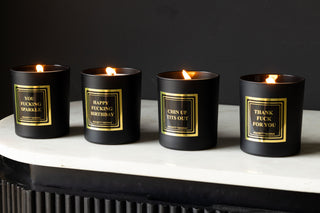 Four scented candles in a row on a white marble console table. The candles are black with gold labels.