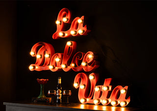 A large fairground-style statement light spelling out the words 'la dolce vita' styled on a wall in the dark above a sideboard with a vase, books, bottle and some wine glasses.