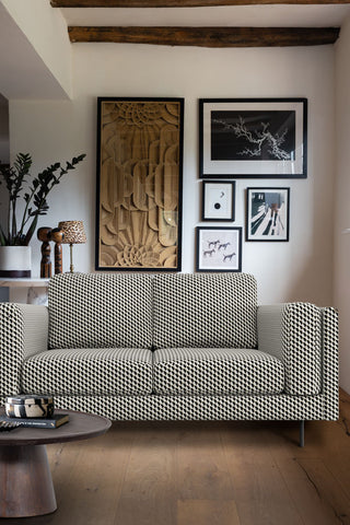 The Grace Medium Sofa In Geo Glam Woven Sand & Black styled in a living room next to two tables styled with various home accessories and several art prints on the wall behind.