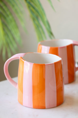 Two Pink & Orange Stripe Mugs displayed on a table with a plant in the background.