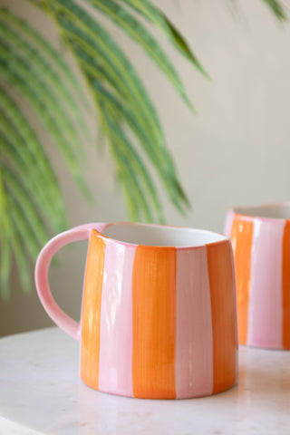 The Pink & Orange Stripe Mug displayed on a table, with another mug and plant in the background.